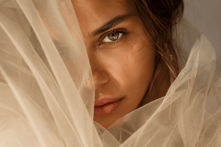 Close-up portret van een vrouw met golvend haar, half gesluierd door doorschijnende tule, in warm zonlicht