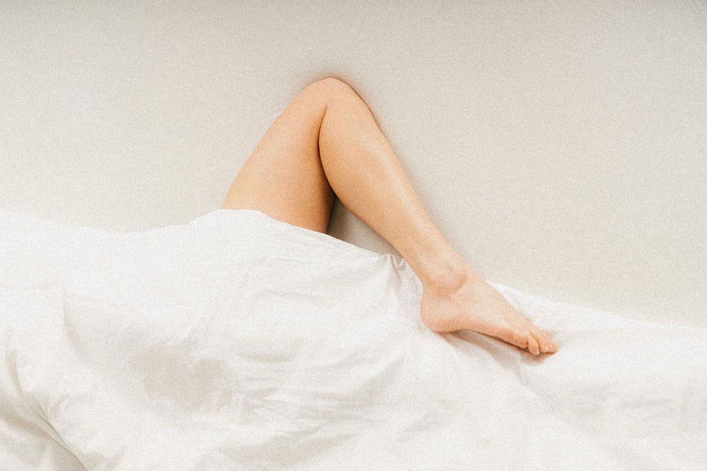 Vrouw ligt in een wit bed met alleen haar benen zichtbaar boven het dekbed tegen een lichte muur, ontspannen sfeer in de slaapkamer.