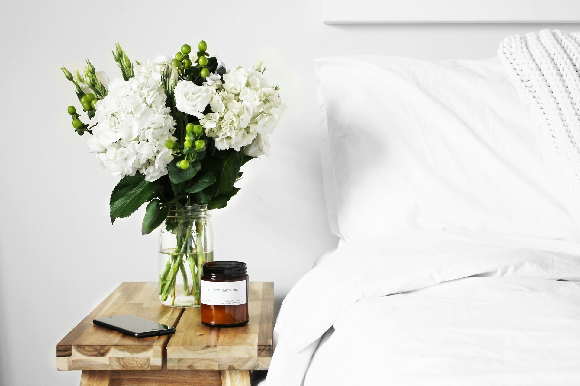een witte kamer met een bed en op het nachtkastje staat een bos bloemen een een verzorgingsproduct