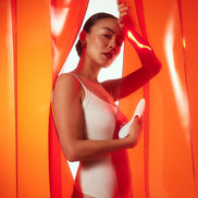 Model in witte body houdt MONA Spectra vast tussen oranje verlichte gordijnen