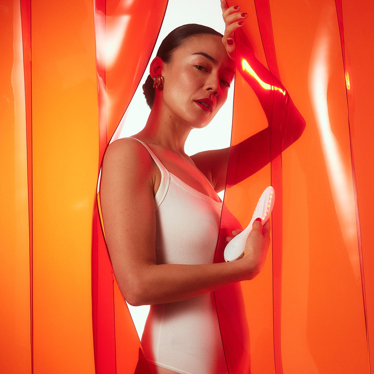 Model in witte body houdt MONA Spectra vast tussen oranje verlichte gordijnen