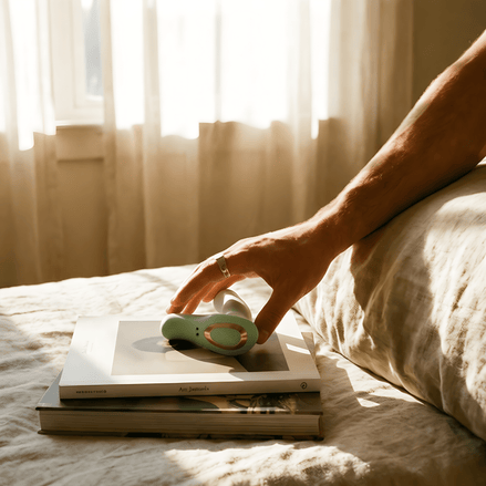 Saliegroene Odes Ombre prostaatvibrator op boeken in warm daglicht, vastgepakt door een hand op bed