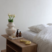 Odes Within strapless strap-on vibrator in beige op houten nachtkastje naast bed, bloemen en verzorgingsproducten