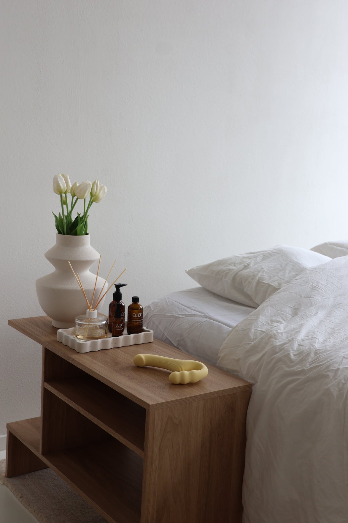 Odes Within strapless strap-on vibrator in beige op houten nachtkastje naast bed, bloemen en verzorgingsproducten