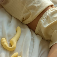 Beige Odes Within strapless strap-on vibrator op wit beddengoed naast een zachte lifestyle setting met model