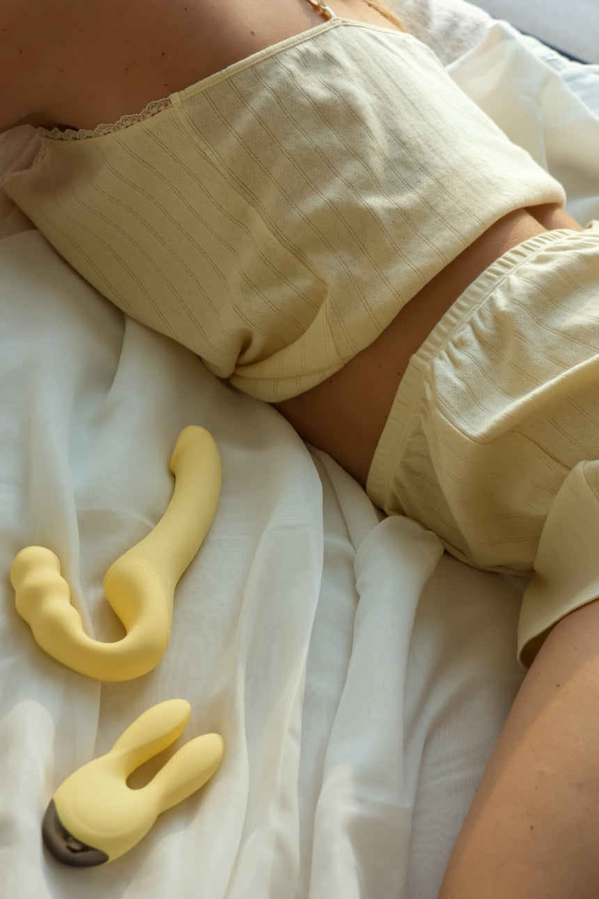 Beige Odes Within strapless strap-on vibrator op wit beddengoed naast een zachte lifestyle setting met model