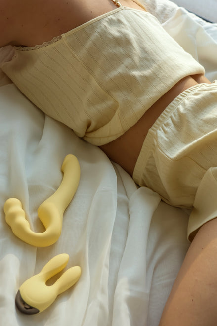 Beige Odes Within strapless strap-on vibrator op wit beddengoed naast een zachte lifestyle setting met model