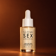 Slow Sex by Bijoux Indiscrets Hair and Skin Shimmer Dry Oil 30 ml, glazen pipetflesje met goudglinsterende droge olie en witte druppelaar tegen een warme koperbruine achtergrond