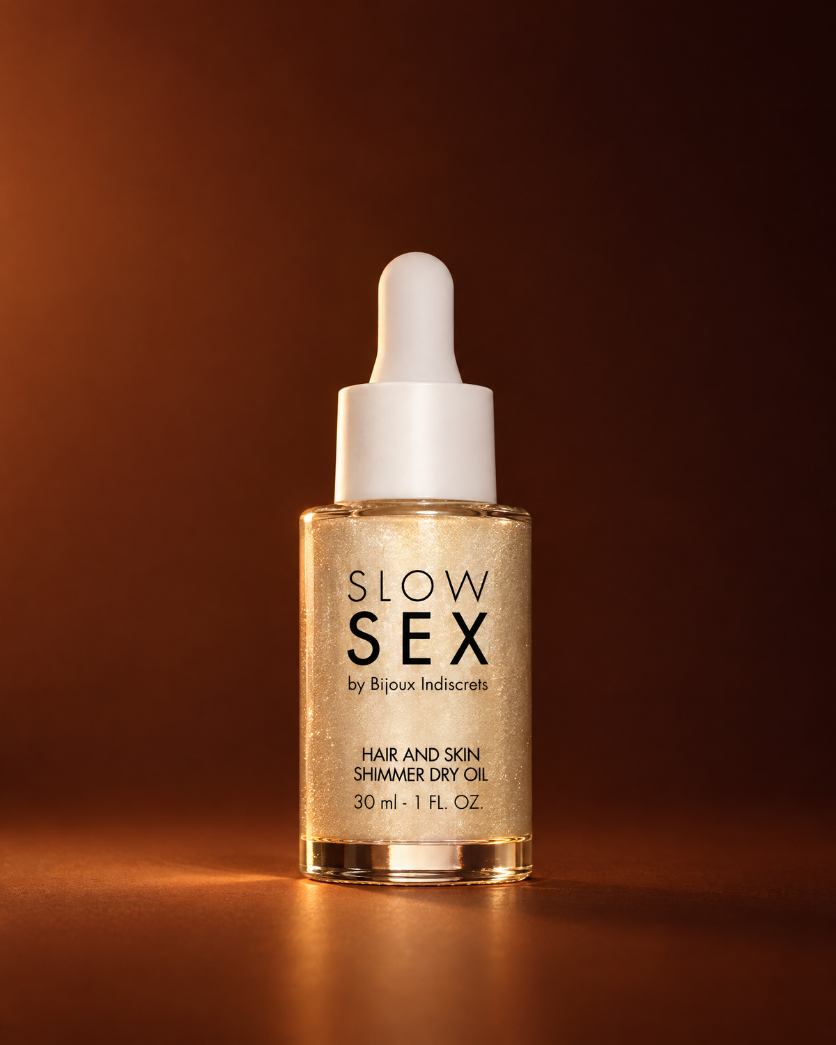 Slow Sex by Bijoux Indiscrets Hair and Skin Shimmer Dry Oil 30 ml, glazen pipetflesje met goudglinsterende droge olie en witte druppelaar tegen een warme koperbruine achtergrond